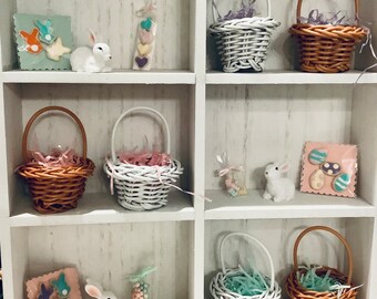 May include: A white wooden shelf displays Easter-themed decorations. Wicker baskets, white ceramic bunnies, and pastel-colored candies are arranged on the shelves. Decorative cards with bunny and egg designs add to the festive display.