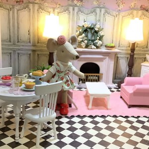 May include: A dollhouse scene with a white table and chairs, a pink armchair, a white coffee table, and a mouse doll wearing a red dress. The scene is set in a dollhouse living room with a fireplace and a wreath on the wall.