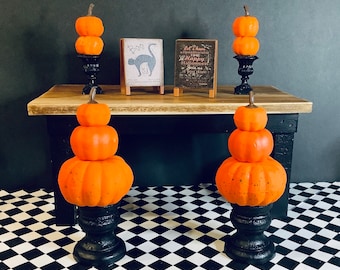 May include: Three tiered pumpkin centerpieces with black stands. Each centerpiece is made of three orange pumpkins stacked on top of each other. The pumpkins are decorated with black and white polka dots. The centerpieces are sitting on a black and white checkered surface. The text on the sign reads "Let's have a spooktacular time Happy Halloween Join us if you dare".
