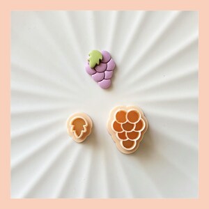 May include: Three cookie cutters, one shaped like a bunch of grapes, one shaped like a single grape, and one shaped like a leaf. The cookie cutters are made of a light pink plastic.