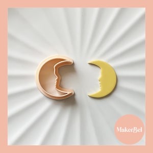 May include: Two crescent moon cookie cutters, one is white plastic and the other is yellow plastic. The cutters are on a white plate with a sunburst pattern.