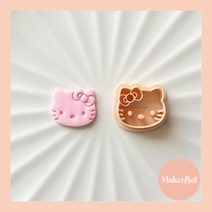 May include: Pink and beige plastic cookie cutters shaped like Hello Kitty. The cutters are on a white surface with a MakerBel logo in the bottom right corner.