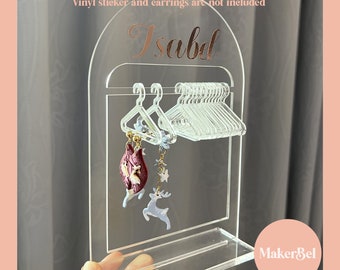 May include: Clear acrylic earring display stand with a small shelf and a row of miniature hangers. The stand has the name "Isabel" written on it in gold lettering. The text "Vinyl sticker and earrings are not included" is written at the top of the stand. The stand is labeled "MakerBel" at the bottom.