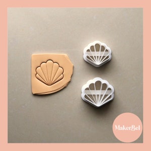 May include: Two white plastic cookie cutters shaped like seashells. The cutters are on a light brown surface. One cutter is on top of a piece of light brown clay, showing the imprint of the cutter.