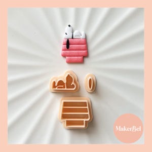 May include: A set of four cookie cutters featuring a dog house, a dog sleeping on top of the house, a cloud, and a circle. The cutters are made of a light beige plastic.