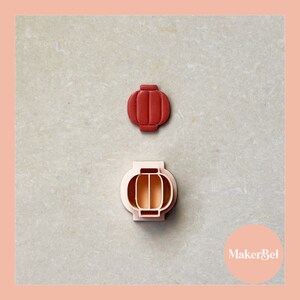 May include: Two red and white plastic cookie cutters shaped like lanterns. The cutters are on a light grey surface. The text "MakerBel" is visible in the bottom right corner of the image.