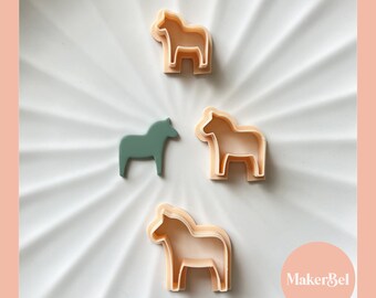 May include: Four cookie cutters in the shape of a horse, three are light peach in color and one is a darker green. The cookie cutters are on a white surface with a swirling pattern. The text "MakerBel" is in the bottom right corner of the image.