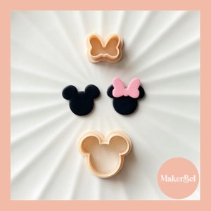 May include: Three plastic cookie cutters in the shape of Mickey Mouse. The cutters are beige and feature a black Mickey Mouse head with large ears and a pink bow. The text "MakerBel" is visible in the lower right corner of the image.