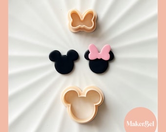 May include: Three plastic cookie cutters in the shape of Mickey Mouse. The cutters are beige and feature a black Mickey Mouse head with large ears and a pink bow. The text "MakerBel" is visible in the lower right corner of the image.