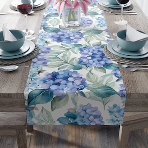 May include: A white table runner with a watercolor floral design featuring blue hydrangeas and green leaves. The runner is placed on a wooden table with two place settings on each side.