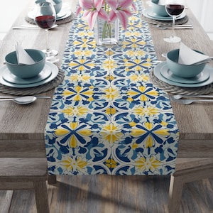Puede incluir: Un camino de mesa azul y amarillo con un diseño floral. El camino de mesa está sobre una mesa de madera con dos cubiertos a cada lado.