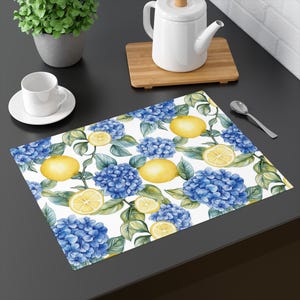 Puede incluir: Salvamanteles blanco con un patrón de limones y hortensias. El salvamanteles presenta flores de hortensia azules y limones amarillos sobre un fondo blanco.
