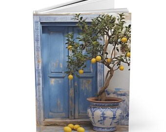 May include: A journal featuring a photograph of a blue door with peeling paint, a potted lemon tree with yellow lemons, and a decorative blue and white pot. Several lemons are on the ground in front of the pot.