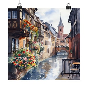 May include: Watercolor painting of a European village with a canal running through it. The buildings are old and have a traditional style, with half-timbered facades and red tile roofs. There are flowers growing on the balconies and along the canal banks. A bridge crosses the canal in the background.