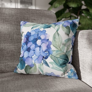 May include: A white throw pillow with a watercolor design of blue and purple hydrangeas and green leaves.