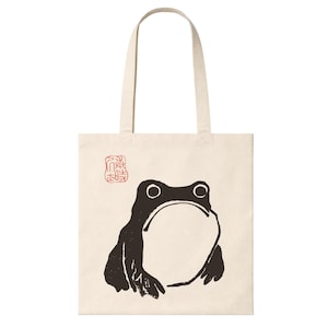 May include: A cream-coloured canvas tote bag featuring a black graphic illustration of a frog in a seated position. Above the frog, there is a small, square red stamp with intricate characters. The bag has two long shoulder straps at the top.