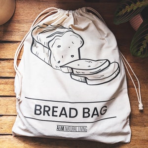 May include: White cotton bread bag with black drawstring and a black and white illustration of bread on the front. The bag has the text "BREAD BAG" and "A&M NATURAL LIVING" printed on the front.