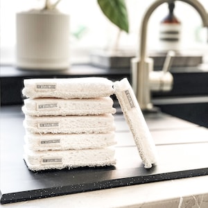 May include: A stack of six white loofah sponges with a black label that reads "AM NATURAL LIVING" on each sponge.