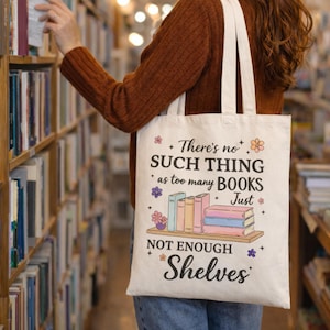 Puede incluir: Un bolso tote crema con el texto "There's no such thing as too many books, just not enough shelves." La bolsa presenta un gr&aacute;fico de libros en un estante, con detalles florales y de estrellas. La bolsa se lleva en una biblioteca.