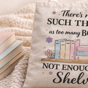 Puede incluir: Un bolso de mano de lona crema con el texto "There's no such thing as too many books, just not enough shelves". El bolso presenta un gr&aacute;fico de libros en una estanter&iacute;a. Tres libros est&aacute;n apilados a la izquierda.