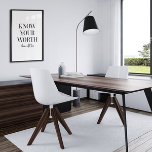 May include: A modern home office featuring a wooden desk and two white chairs. A framed print with the text "KNOW YOUR WORTH" is displayed on the wall. A black floor lamp with a white shade stands in the corner.