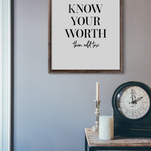 May include: A framed print with the text "KNOW YOUR WORTH then add tax" on a white background. The frame is brown wood. The print is hanging on a light grey wall.