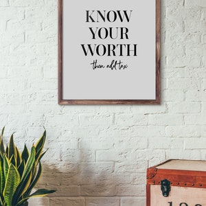 May include: A framed print with a white background and black text that reads "KNOW YOUR WORTH then add tax". The print is hanging on a white brick wall. A potted plant and a wooden chest with numbers on the front are in the foreground.