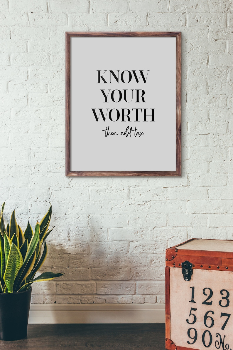 May include: A framed print with a white background and black text that reads "KNOW YOUR WORTH then add tax". The print is hanging on a white brick wall. A potted plant and a wooden chest with numbers on the front are in the foreground.