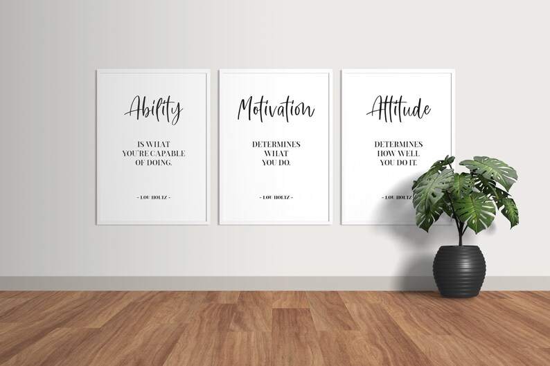 May include: Three framed motivational posters with white backgrounds and black text. The posters are hung on a white wall. The text on the posters reads: "Ability is what you're capable of doing", "Motivation determines what you do", and "Attitude determines how well you do it". The posters are attributed to Lou Holtz. A potted green plant sits on the wooden floor in front of the posters.