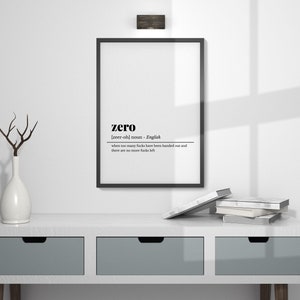 May include: A framed print with the definition of "zero" in black text. The print hangs above a white console table with three gray drawers. A white vase with a branch and a stack of books are on the table.
