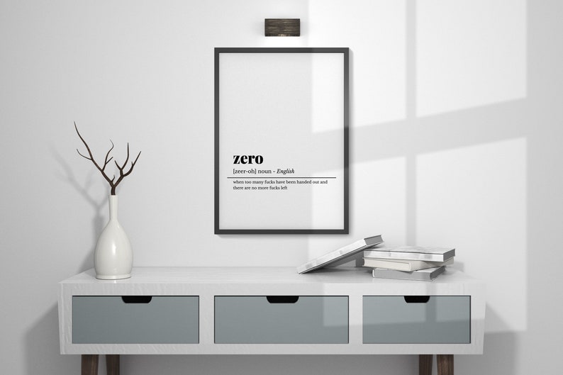 May include: A framed print with the definition of "zero" in black text. The print hangs above a white console table with three gray drawers. A white vase with a branch and a stack of books are on the table.