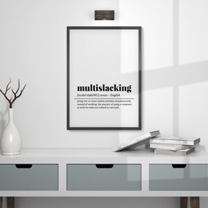 May include: A black and white framed print with the definition of the word "multislacking" in English. The print is hanging on a white wall above a white dresser with two drawers. There are books stacked on the dresser.