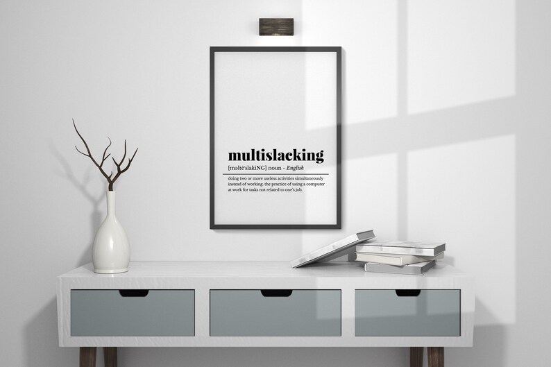 May include: A black and white framed print with the definition of the word "multislacking" in English. The print is hanging on a white wall above a white dresser with two drawers. There are books stacked on the dresser.
