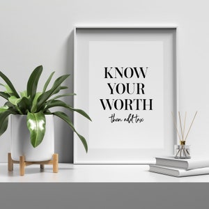 May include: A white framed print with the text "KNOW YOUR WORTH then add tax" in black lettering on a white background. The print is sitting on a white surface with a potted plant and a diffuser to the left and a stack of books to the right.