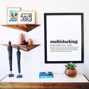 May include: A black framed print with the definition of the word "multislacking" in English. The print is hanging on a white wall above a wooden dresser with two wooden shelves. The shelves are decorated with two wooden statues, two terracotta vases, and a black vase. A small potted succulent plant sits on the dresser.
