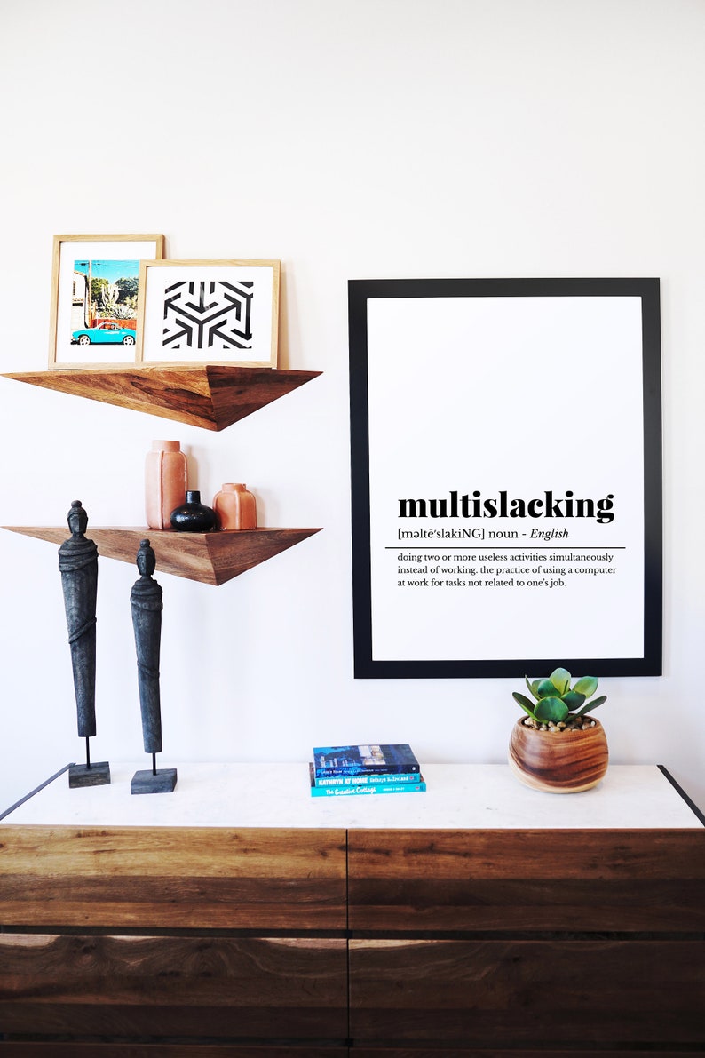 May include: A black framed print with the definition of the word "multislacking" in English. The print is hanging on a white wall above a wooden dresser with two wooden shelves. The shelves are decorated with two wooden statues, two terracotta vases, and a black vase. A small potted succulent plant sits on the dresser.