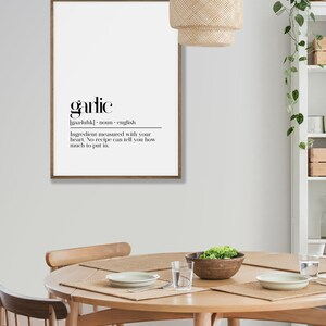 May include: A framed black and white print with the word "garlic" in bold font. The definition of garlic is written below in a smaller font. The print is hanging on a white wall above a wooden table with chairs.
