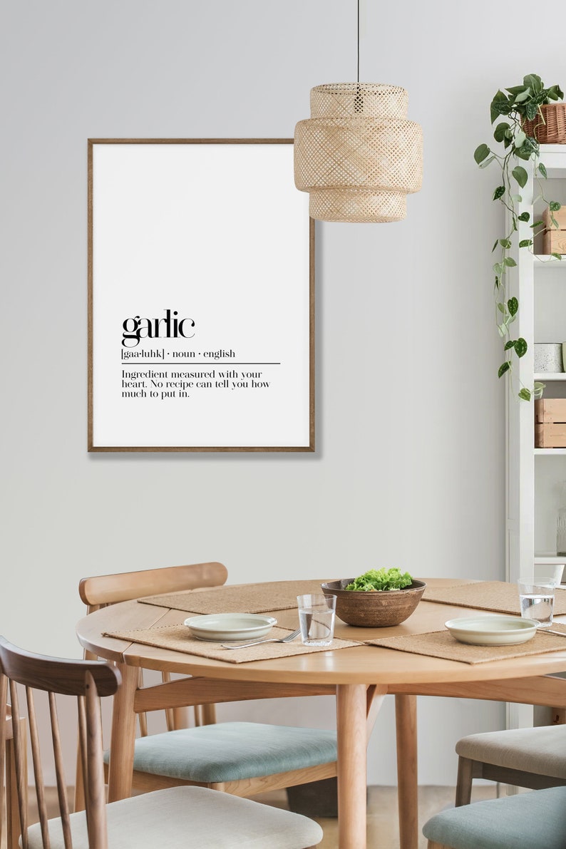 May include: A framed black and white print with the word "garlic" in bold font. The definition of garlic is written below in a smaller font. The print is hanging on a white wall above a wooden table with chairs.