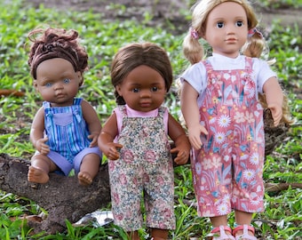 Op de afbeelding: Drie poppen die overalls dragen. De pop links heeft bruin, krullend haar en blauwe ogen. De pop in het midden heeft bruin haar en bruine ogen. De pop rechts heeft blond haar en blauwe ogen. Alle drie de poppen dragen overalls met bloemen.