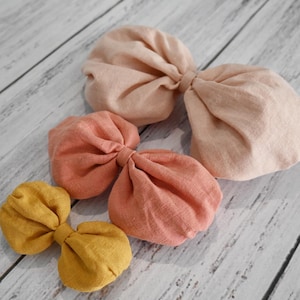 May include: Four fabric bows in different sizes and colours. The largest bow is a light peach colour, the next largest is a coral colour, the next is a darker coral colour, and the smallest bow is a mustard yellow colour.
