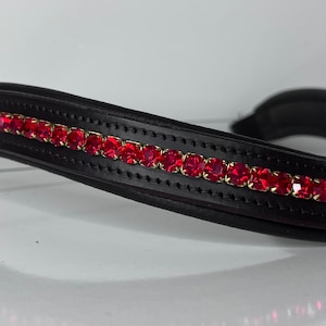 May include: Black leather horse bridle browband with a row of vibrant red square-cut rhinestones set in gold-tone metal. The browband is curved and padded for comfort, with a polished finish.