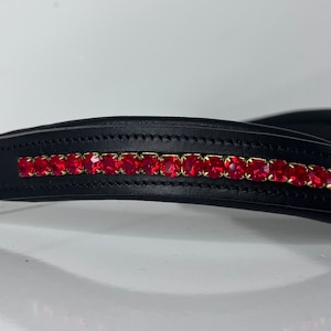 May include: Black leather horse bridle with a row of vibrant red rhinestones set in gold-colored metal. The bridle is designed with a curved shape and is likely used for equestrian activities.
