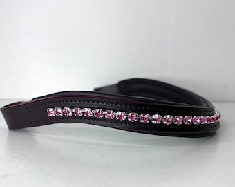 May include: A dark brown leather horse browband, adorned with a line of pink rhinestones. The browband is curved and padded. The background is white.