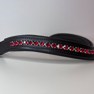 May include: Black leather horse bridle with a row of red rhinestones. The bridle is curved and has a stitched design. The red rhinestones are set in a silver-colored metal. The background is white.