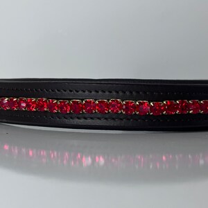 May include: Black leather browband with a row of vibrant red rhinestones. The browband features a rectangular shape and is designed for equestrian use. The red stones are set in a gold-colored metal.