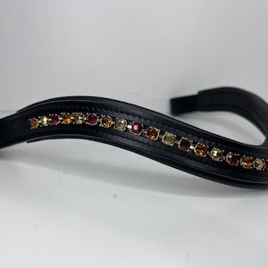 May include: Black leather horse bridle browband with a curved design. The browband is adorned with a row of colorful, sparkling rhinestones in shades of red, gold, and amber. The browband is designed for equestrian use.