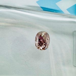 May include: An oval-shaped, pink diamond with a complex internal structure. The gem is set against a light blue and white background, creating a contrast that highlights its color and brilliance. The diamond's facets reflect light, giving it a sparkling appearance.