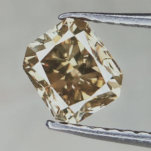 May include: A radiant-cut, light brown diamond held by a pair of silver tweezers. The gemstone has multiple facets that reflect light, creating a sparkling effect. The diamond's shape is a modified square with cut corners, showcasing its brilliance.