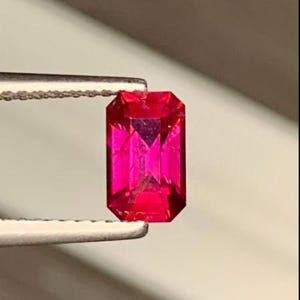 May include: A vibrant, rectangular-cut, hot pink gemstone is held by tweezers. The gem's facets reflect light, showcasing its clarity and brilliance. This gemstone is a striking example of fine jewelry.