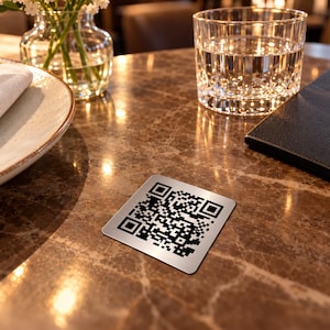 Op de afbeelding: Een vierkante onderzetter van geborsteld zilverkleurig metaal met een zwarte QR-code. De onderzetter heeft afgeronde hoeken en een metalen afwerking, met een digitaal patroon met hoog contrast dat is ontworpen om te scannen.